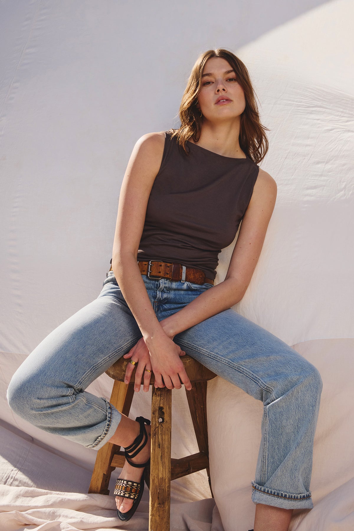 A person sits on a wooden stool, wearing an EMILIA TANK TOP by Velvet by Graham & Spencer and rolled jeans, hands resting gently on their lap against a plain backdrop.-39003862991041