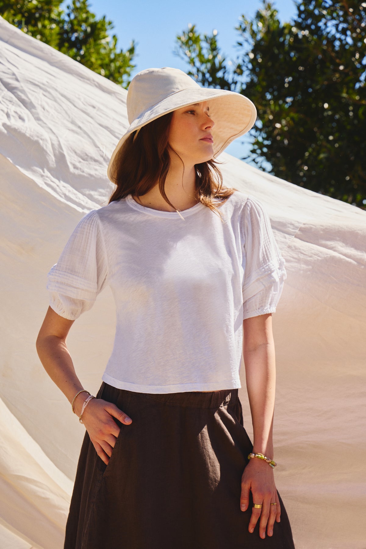 A woman in the HADEN TOP by Velvet by Graham & Spencer and a wide-brimmed hat poses outdoors against a fabric backdrop, with trees and blue sky behind her.-39003864891585