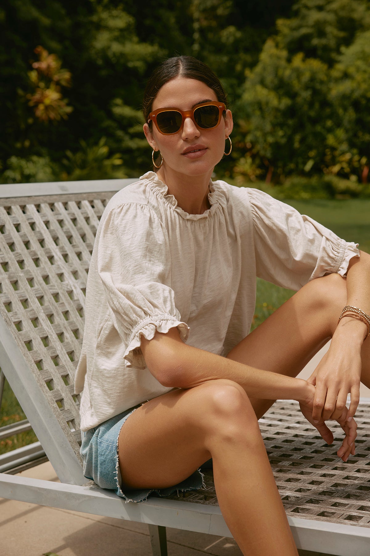   A woman wearing sunglasses,  the CARLENE TEE by Velvet by Graham & Spencer, and denim shorts sits on a metal outdoor chair in a garden, enjoying the relaxed fit of her breathable cotton outfit. 