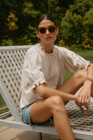 A woman wearing sunglasses,  the CARLENE TEE by Velvet by Graham & Spencer, and denim shorts sits on a metal outdoor chair in a garden, enjoying the relaxed fit of her breathable cotton outfit.