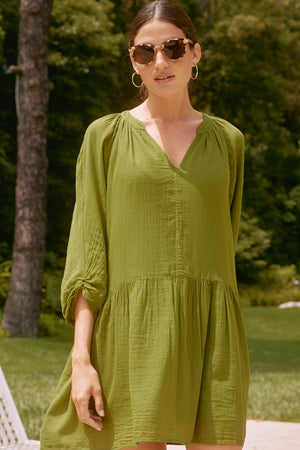 A woman wearing a green SLOAN DRESS by Velvet by Graham & Spencer, made of breathable cotton gauze, and sunglasses stands outdoors with trees in the background.