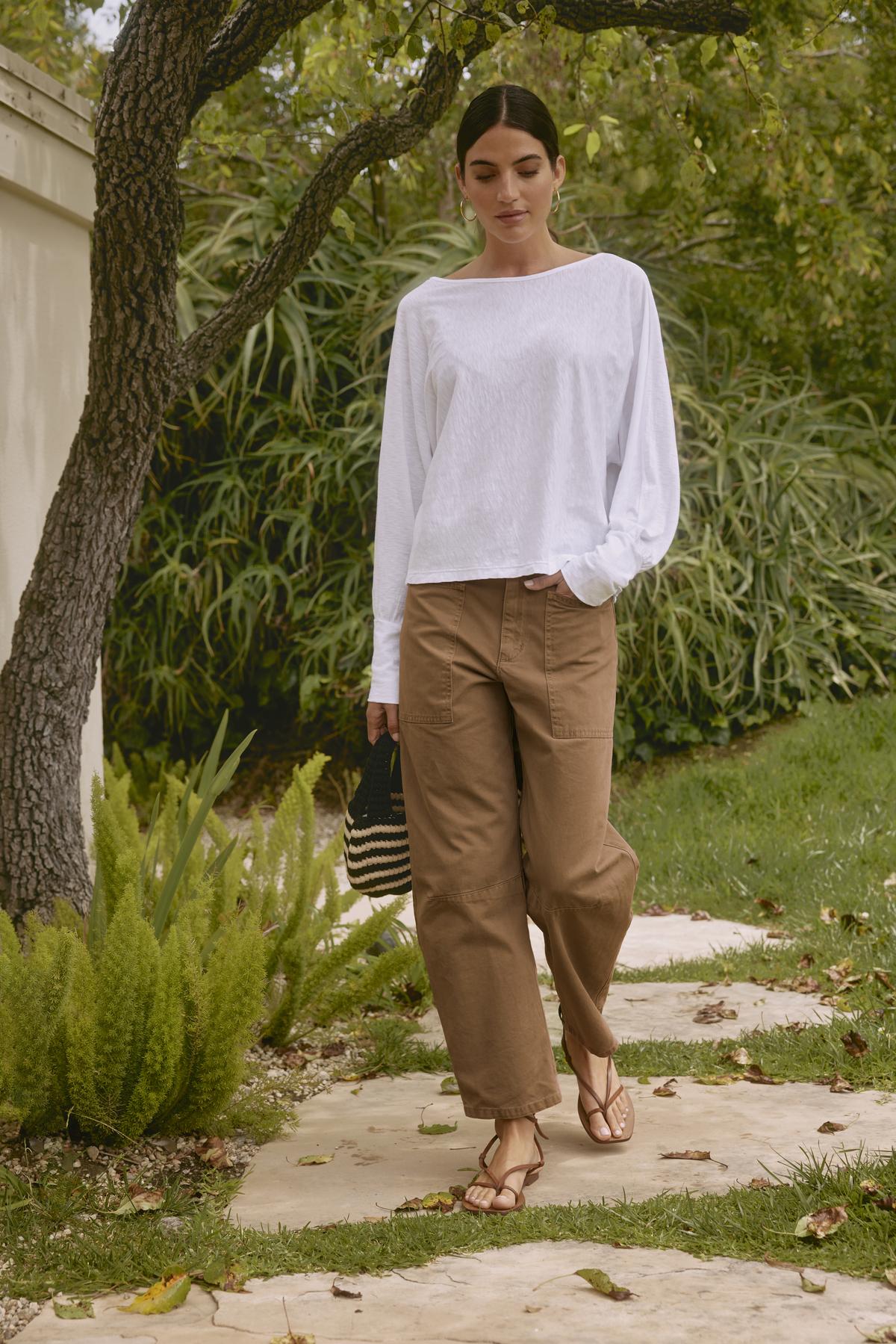   A person wearing a white long-sleeve shirt and the BRYLIE SANDED TWILL UTILITY PANT by Velvet by Graham & Spencer, holding a black straw bag, walking on a stone path in a garden with green foliage. 