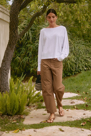 A person wearing a white long-sleeve shirt and the BRYLIE SANDED TWILL UTILITY PANT by Velvet by Graham & Spencer, holding a black straw bag, walking on a stone path in a garden with green foliage.