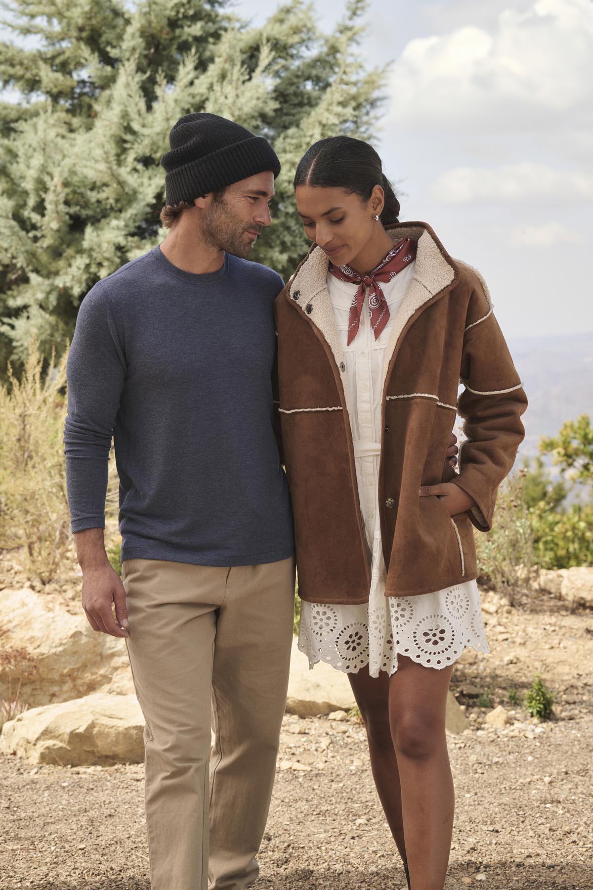 A man in a beanie and a Velvet by Graham & Spencer BECKER CREW sweater walks with a woman wearing a brown jacket and scarf. They are outdoors near a tree with hills in the background, enjoying the comfort of daily wear.-38282364289217