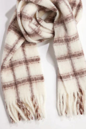 The WARM WINDOW PLAID SCARF by Velvet by Graham & Spencer, featuring a cream base with a brown plaid pattern and fringe ends, is displayed on a flat surface.