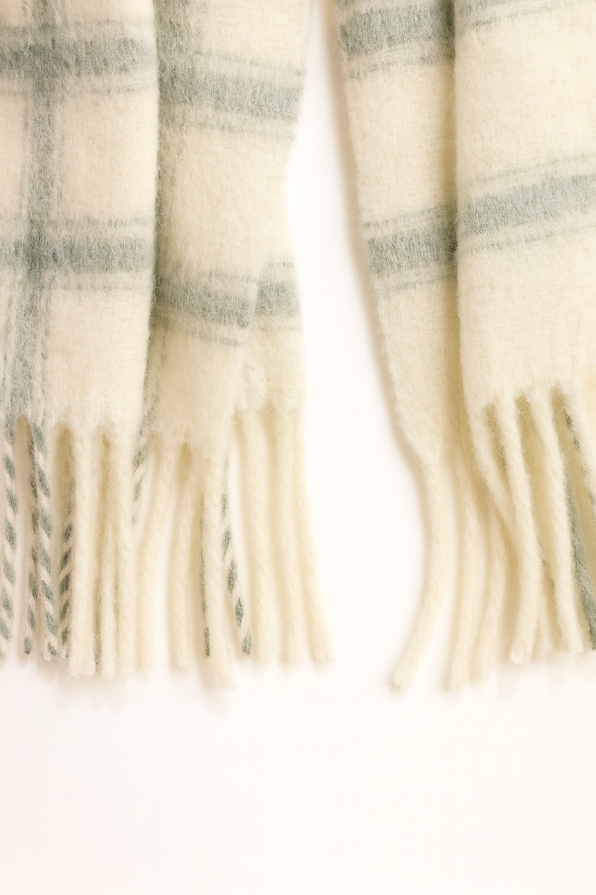 The WARM WINDOW PLAID SCARF by Velvet by Graham & Spencer features a cream base with light green plaid and fringed ends, displayed on a white surface.