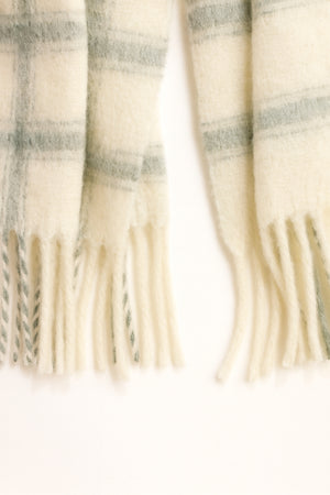 The WARM WINDOW PLAID SCARF by Velvet by Graham & Spencer features a cream base with light green plaid and fringed ends, displayed on a white surface.