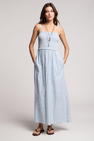 A woman wears the SENARA DRESS by Velvet by Graham & Spencer, a sleeveless maxi with watercolor stripes, adjustable straps, and pockets. She pairs it with sandals and layered necklaces.