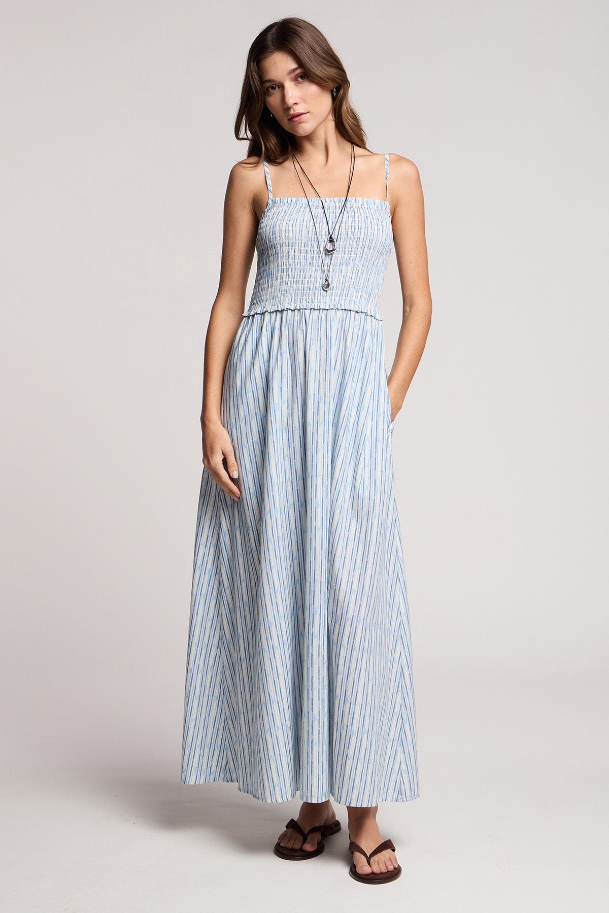 Woman in a studio wears the SENARA DRESS by Velvet by Graham & Spencer, a sleeveless maxi dress with watercolor stripes and pockets, styled with a long necklace and sandals.-41372216099009