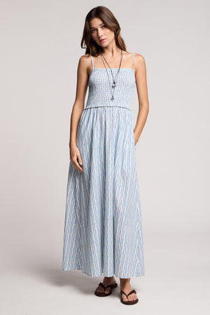 Woman in a studio wears the SENARA DRESS by Velvet by Graham & Spencer, a sleeveless maxi dress with watercolor stripes and pockets, styled with a long necklace and sandals.