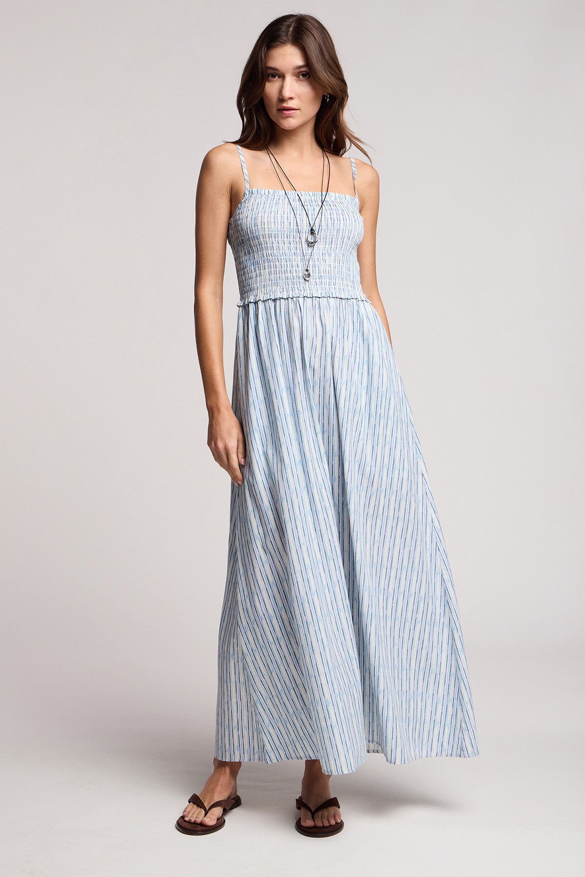 A woman stands in a studio wearing the SENARA DRESS by Velvet by Graham & Spencer—a sleeveless maxi with adjustable straps and watercolor stripes—styled with necklaces and sandals.-41372216131777