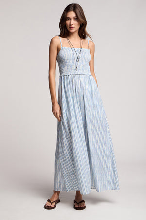 A woman stands in a studio wearing the SENARA DRESS by Velvet by Graham & Spencer—a sleeveless maxi with adjustable straps and watercolor stripes—styled with necklaces and sandals.