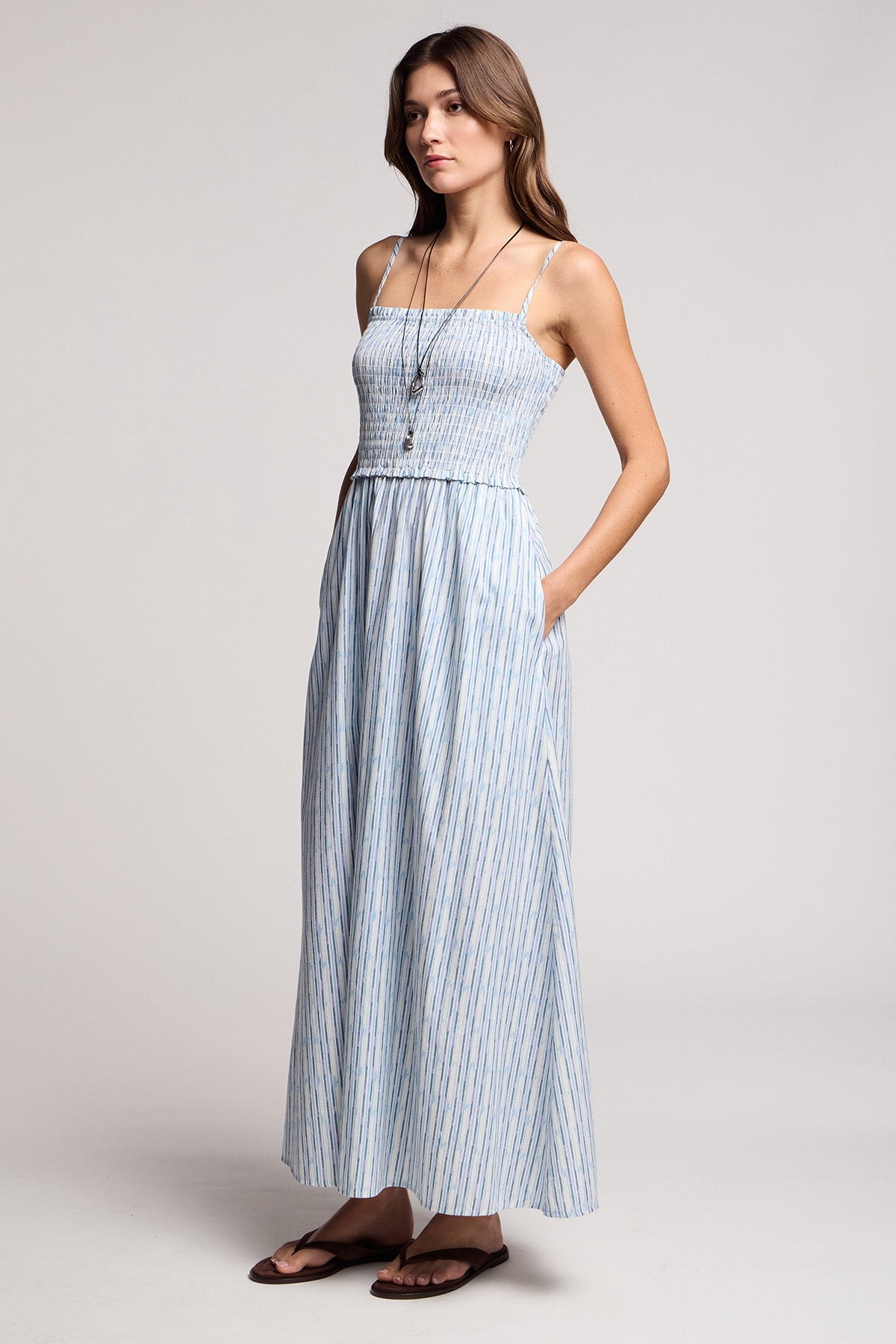 A woman wears the SENARA DRESS by Velvet by Graham & Spencer—a sleeveless, ankle-length watercolor stripe maxi with pockets—paired with a long necklace and brown sandals against a plain background.-41372216164545