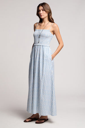A woman wears the SENARA DRESS by Velvet by Graham & Spencer—a sleeveless, ankle-length watercolor stripe maxi with pockets—paired with a long necklace and brown sandals against a plain background.