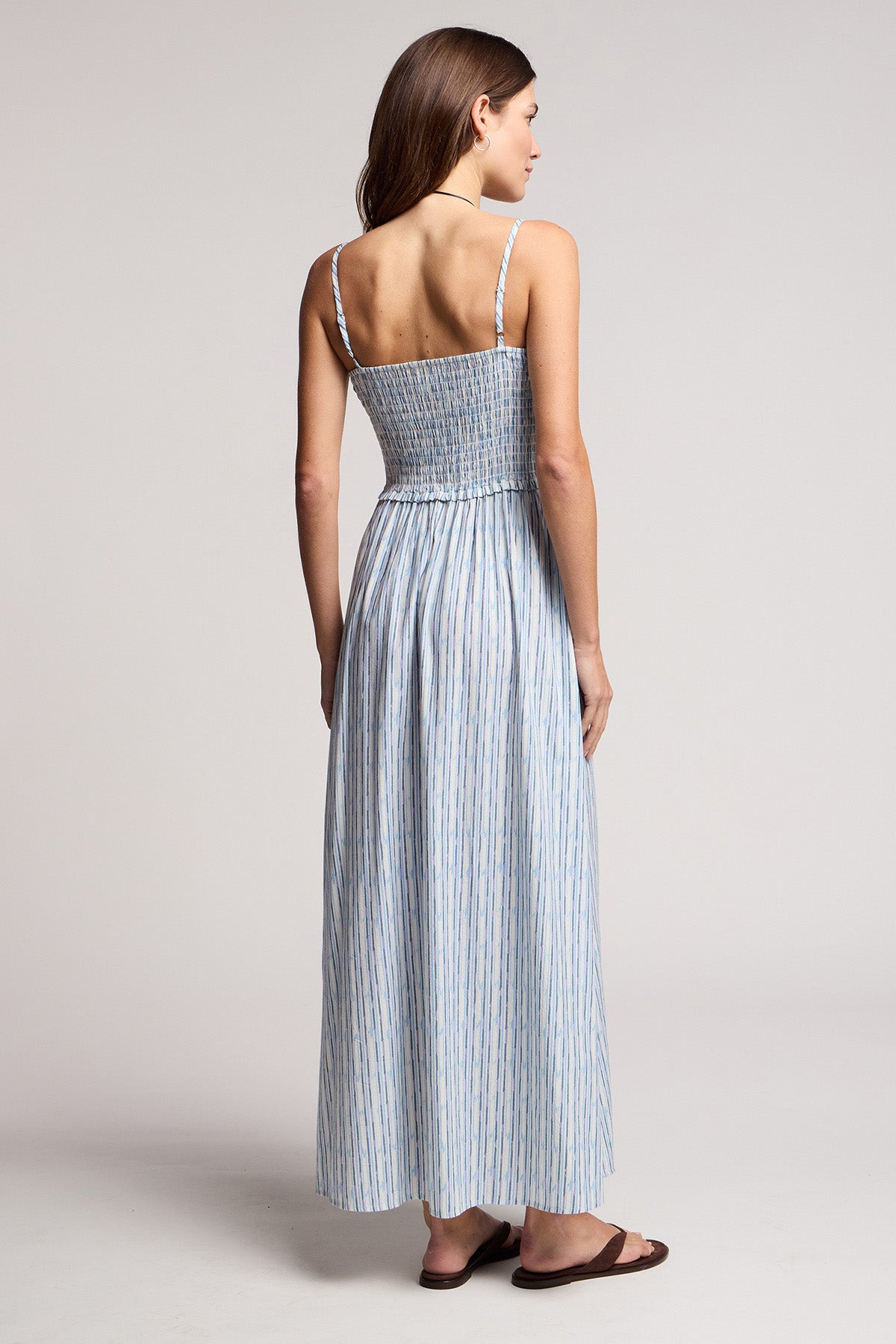 A woman faces away wearing the SENARA DRESS by Velvet by Graham & Spencer, a watercolor stripe maxi with thin adjustable straps, paired with flat sandals.
