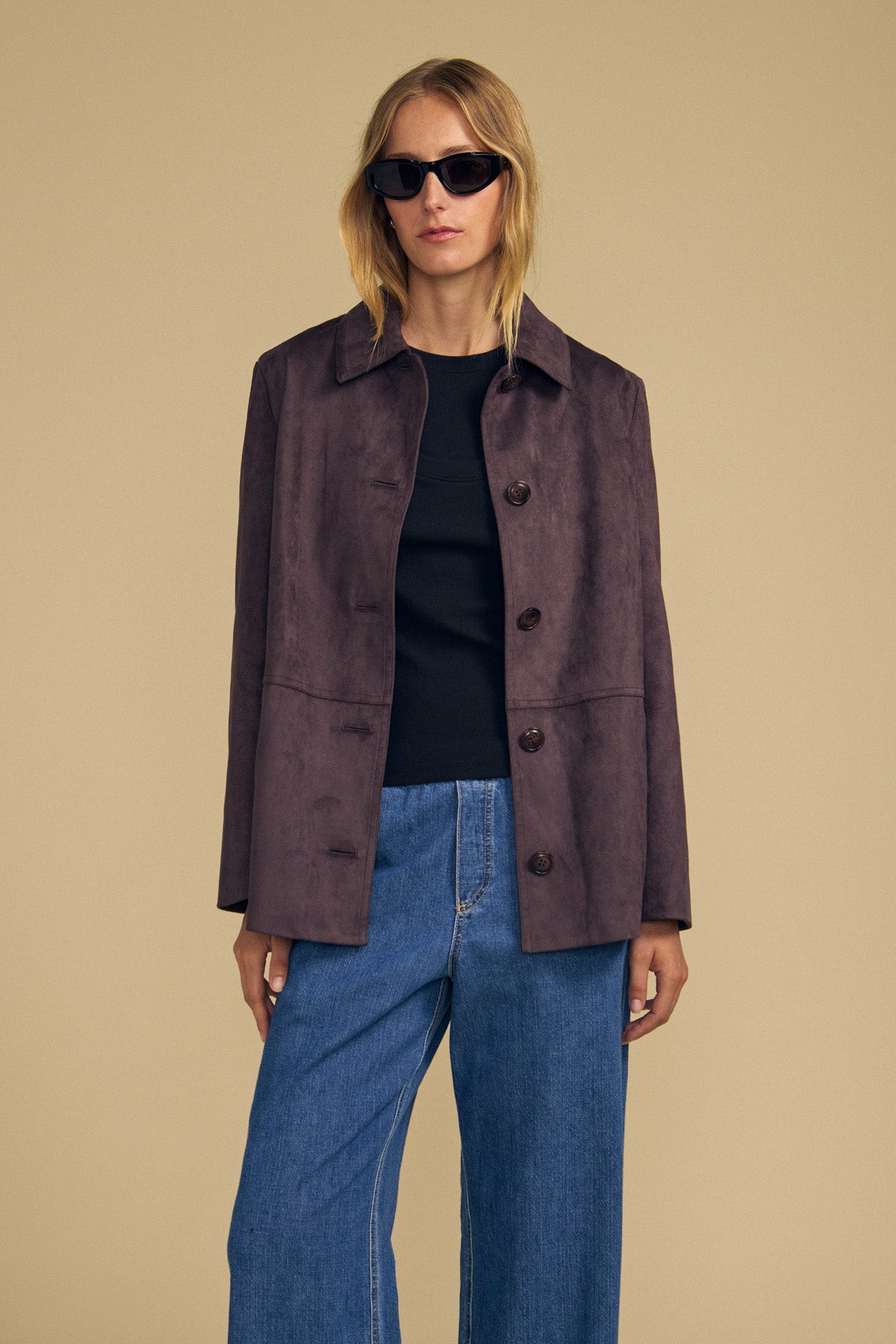 Wearing the WENDOLYN FAUX SUEDE JACKET by Velvet by Graham & Spencer in dark brown over a black shirt and blue wide-leg jeans, a person stands against a beige background, accessorized with sunglasses.
