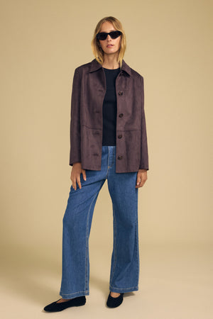 A woman wearing sunglasses, the Velvet by Graham & Spencer WENDOLYN FAUX SUEDE JACKET in dark brown with a classic silhouette, a black top, wide-leg blue jeans, and black shoes stands against a plain beige background.