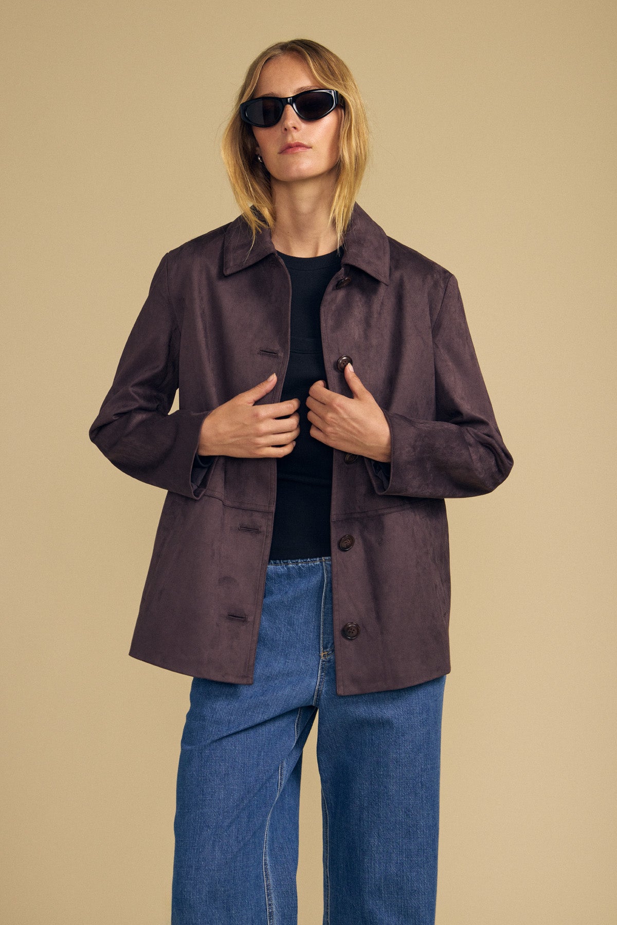 Model wearing sunglasses, styled in the Velvet by Graham & Spencer WENDOLYN FAUX SUEDE JACKET layered over a black top and blue jeans, stands against a plain beige background.-41976525226177