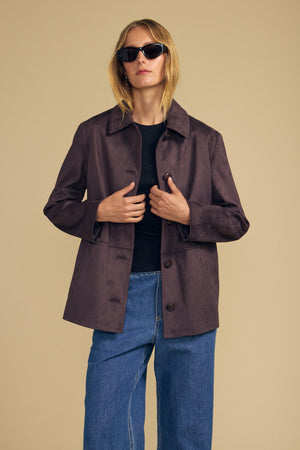 Model wearing sunglasses, styled in the Velvet by Graham & Spencer WENDOLYN FAUX SUEDE JACKET layered over a black top and blue jeans, stands against a plain beige background.