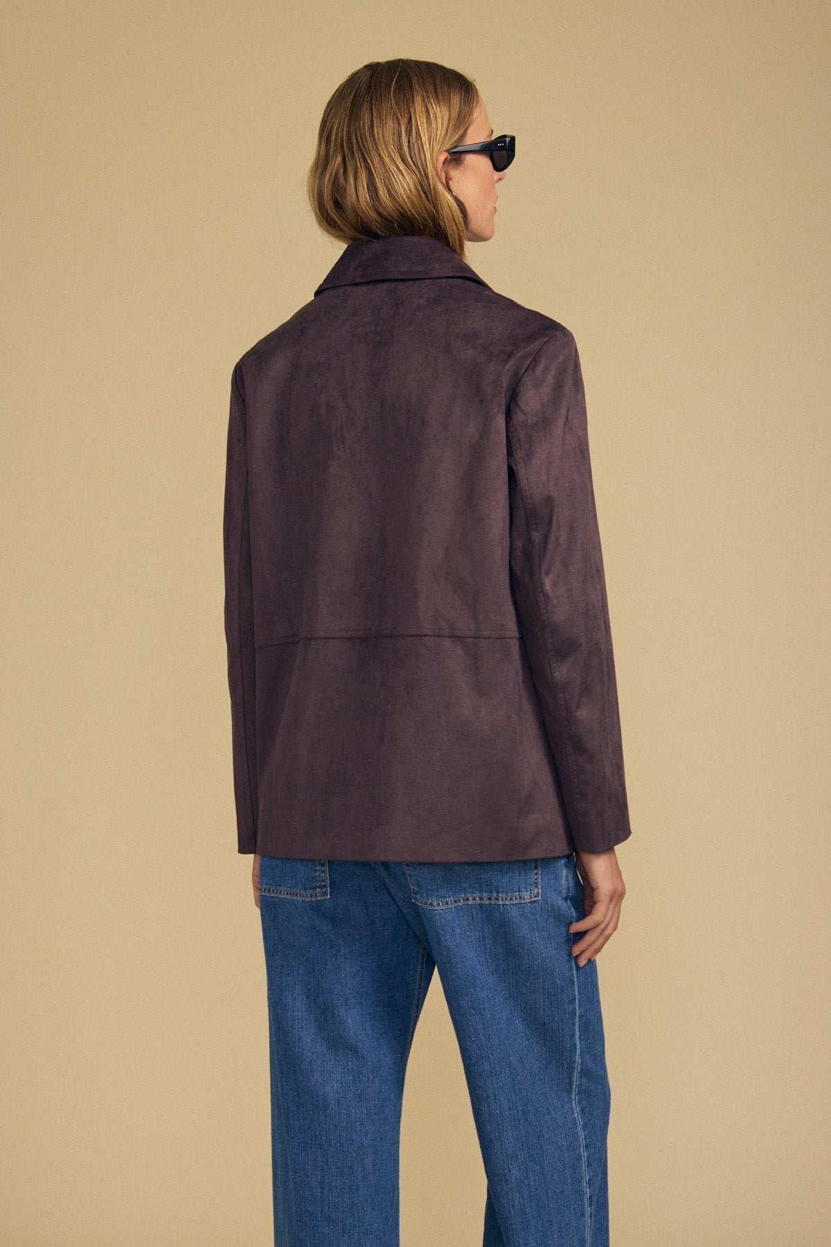 A person stands facing away against a plain beige background, wearing sunglasses, blue jeans, and the WENDOLYN FAUX SUEDE JACKET by Velvet by Graham & Spencer—a button-front jacket with a classic silhouette.