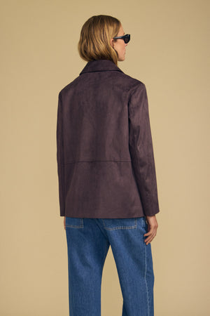 A person stands facing away against a plain beige background, wearing sunglasses, blue jeans, and the WENDOLYN FAUX SUEDE JACKET by Velvet by Graham & Spencer—a button-front jacket with a classic silhouette.