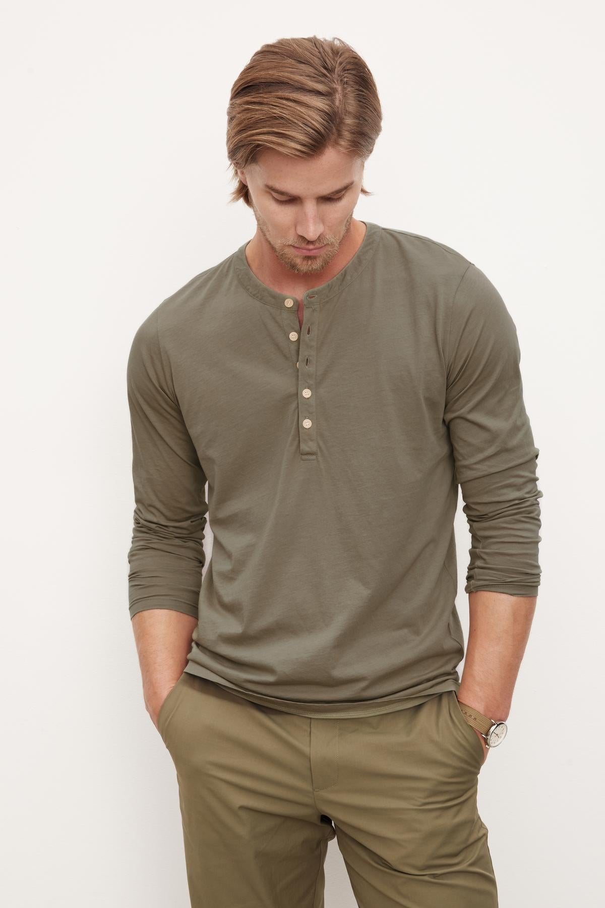   A man in a casual olive green Velvet by Graham & Spencer ALVARO COTTON JERSEY HENLEY shirt and matching pants with a vintage-look, looking down thoughtfully, with a wristwatch visible on his left arm. 