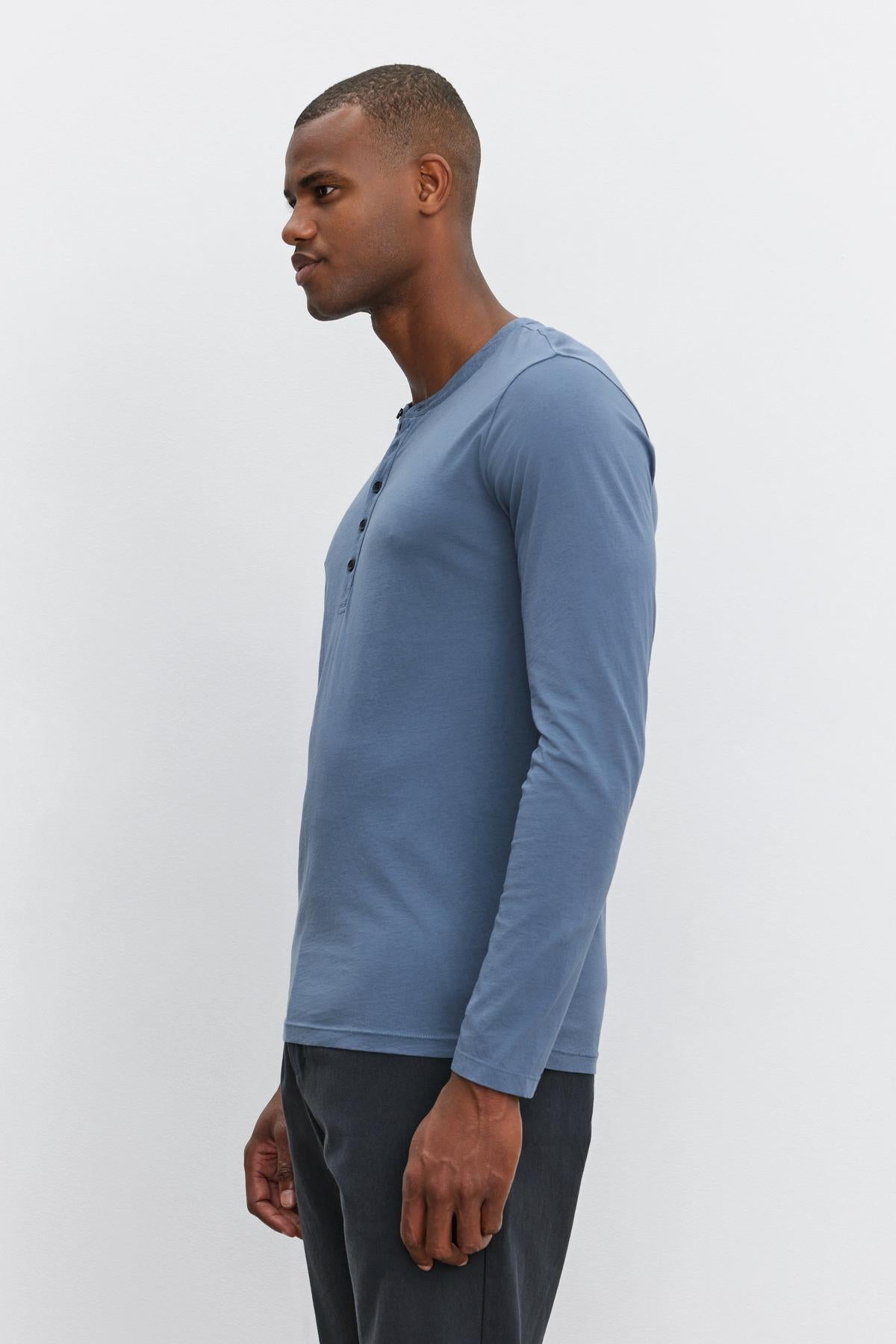   A man stands against a light background, wearing the Velvet by Graham & Spencer ALVARO HENLEY in lightweight blue and dark pants. He is facing left in a side profile view. 