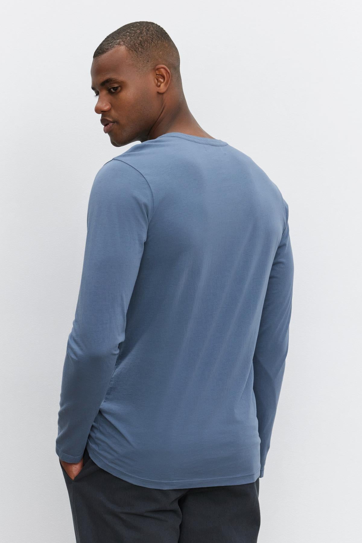   A person wearing the ALVARO HENLEY, a long-sleeved blue lightweight shirt by Velvet by Graham & Spencer, paired with dark pants, is standing with their back to the camera, looking to the side. 