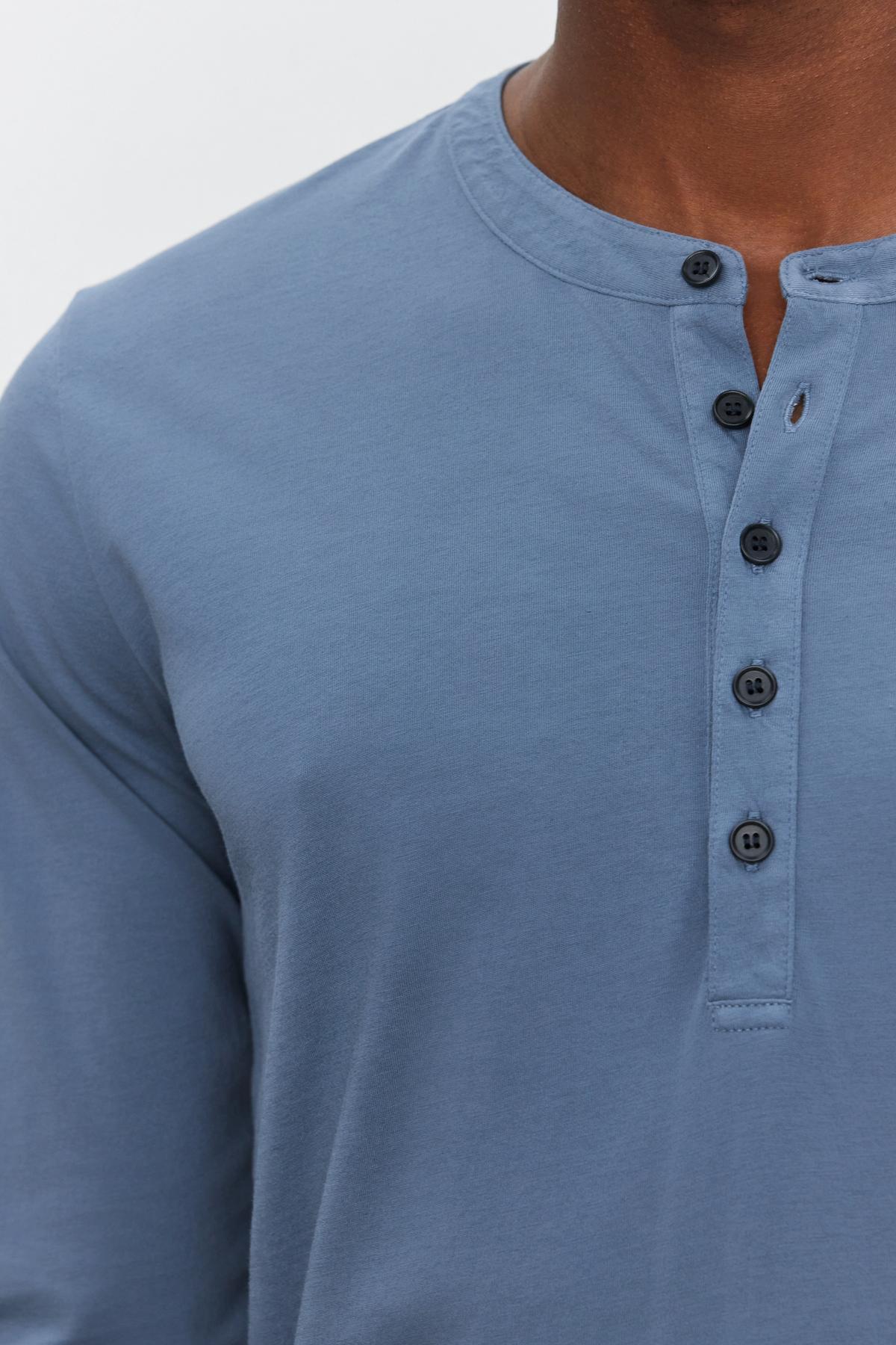 Close-up of a person wearing the soft, lightweight ALVARO HENLEY shirt by Velvet by Graham & Spencer, featuring a light blue color, crew neckline, and multiple black buttons down the front.-37759069847745