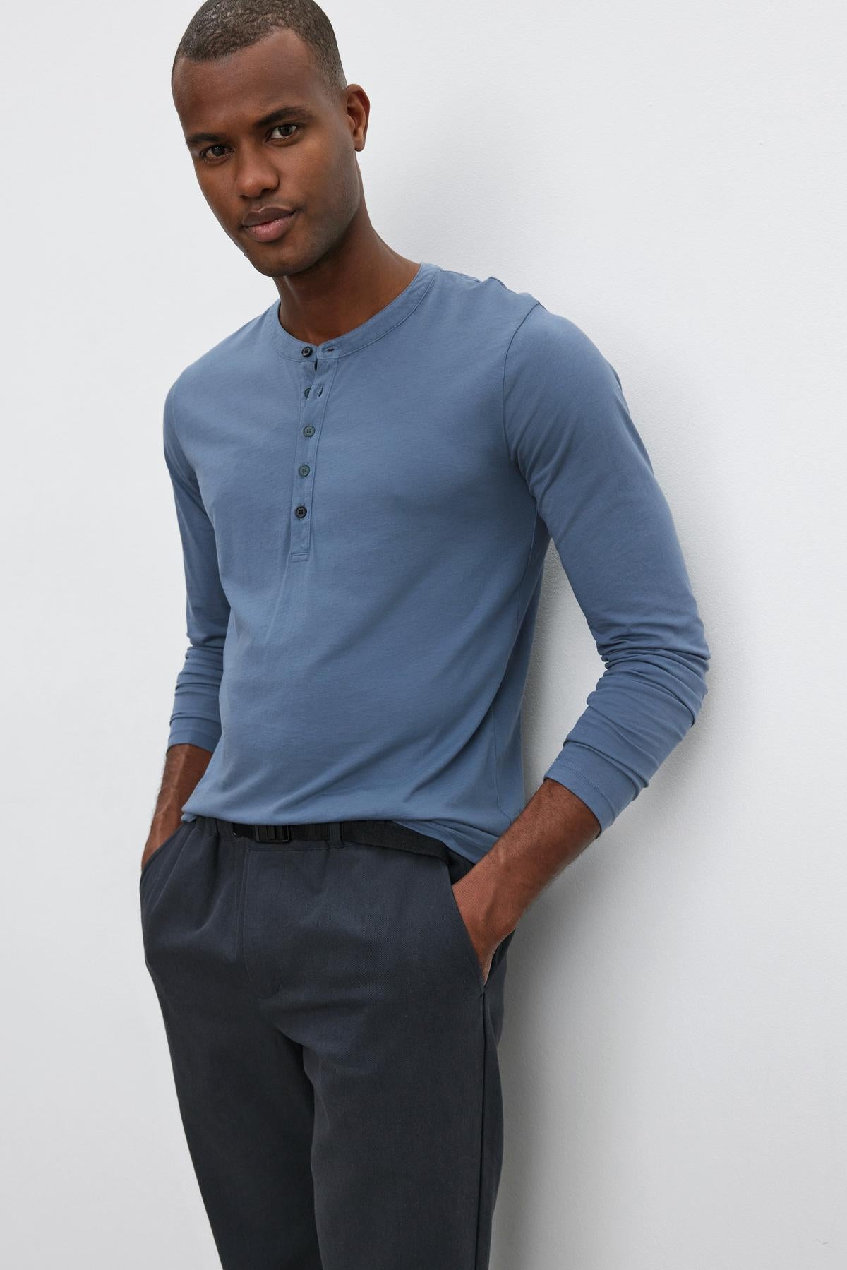 A man with short hair stands casually with hands in pockets, wearing the ALVARO HENLEY by Velvet by Graham & Spencer— a lightweight blue henley shirt that's perfect for layering. He pairs it with dark pants, all set against a plain white background.-37759069749441