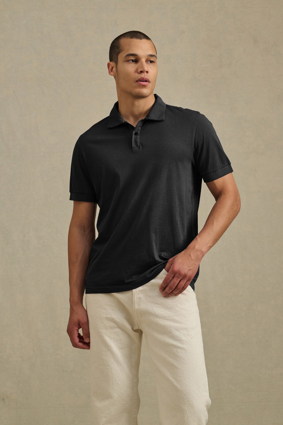 A man wears the ARROW POLO by Velvet by Graham & Spencer in black cotton with off-white pants, standing against a neutral textured background for a classic, effortless casual look.