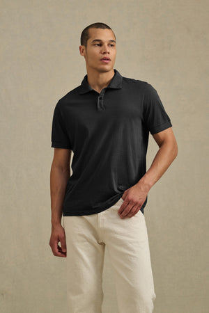 A man wears the ARROW POLO by Velvet by Graham & Spencer in black cotton with off-white pants, standing against a neutral textured background for a classic, effortless casual look.