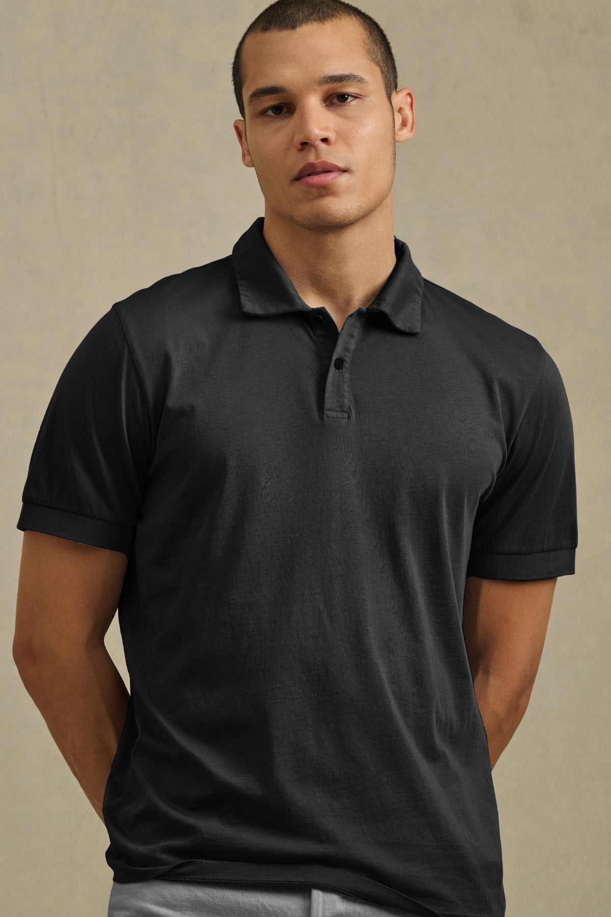 A person in the ARROW POLO by Velvet by Graham & Spencer—a short-sleeve black cotton polo—paired with light gray pants stands against a plain beige background, showcasing timeless style for casual looks.