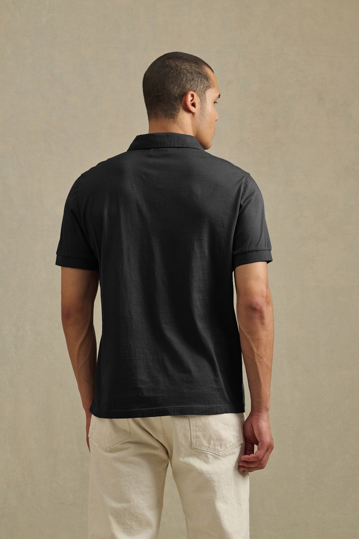 A man faces away from the camera, wearing the ARROW POLO by Velvet by Graham & Spencer, a short-sleeve black casual polo, paired with light-colored pants against a neutral background.-41434440368321