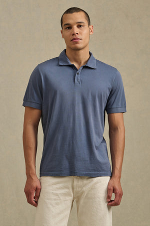 A man faces forward with a neutral expression, wearing the Velvet by Graham & Spencer ARROW POLO, a classic short-sleeve blue cotton shirt, paired with off-white pants against a plain background.