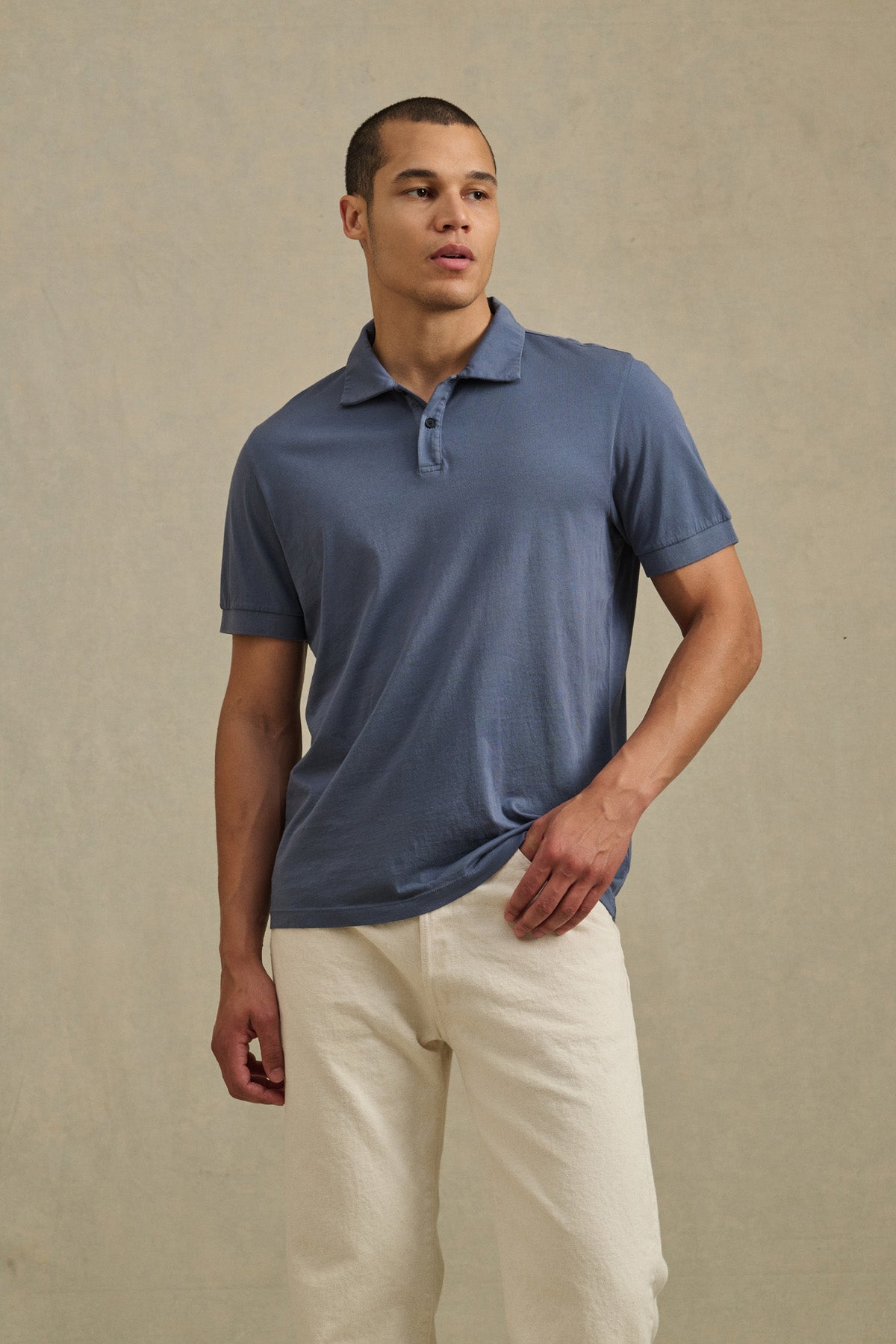 A man stands against a plain beige background wearing the ARROW POLO by Velvet by Graham & Spencer in blue cotton and off-white pants, looking to his left.