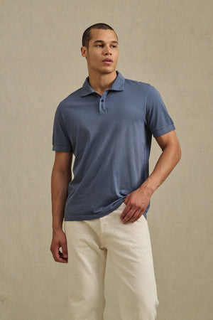 A man stands against a plain beige background wearing the ARROW POLO by Velvet by Graham & Spencer in blue cotton and off-white pants, looking to his left.