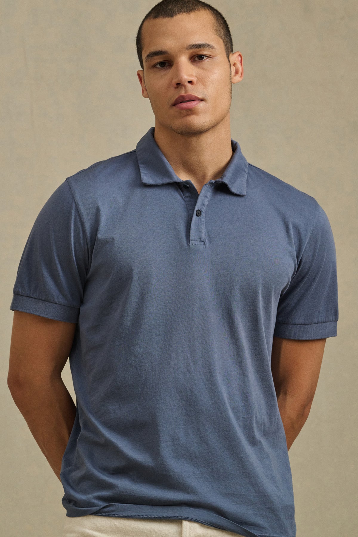 A man wears the ARROW POLO by Velvet by Graham & Spencer, a short-sleeve blue cotton shirt, standing against a beige background and looking directly at the camera for a classic style.-41104666296513