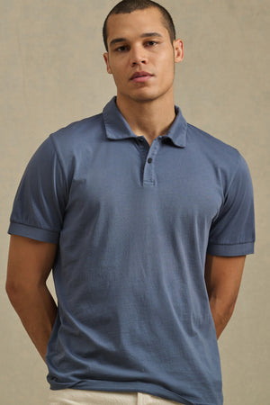 A man wears the ARROW POLO by Velvet by Graham & Spencer, a short-sleeve blue cotton shirt, standing against a beige background and looking directly at the camera for a classic style.