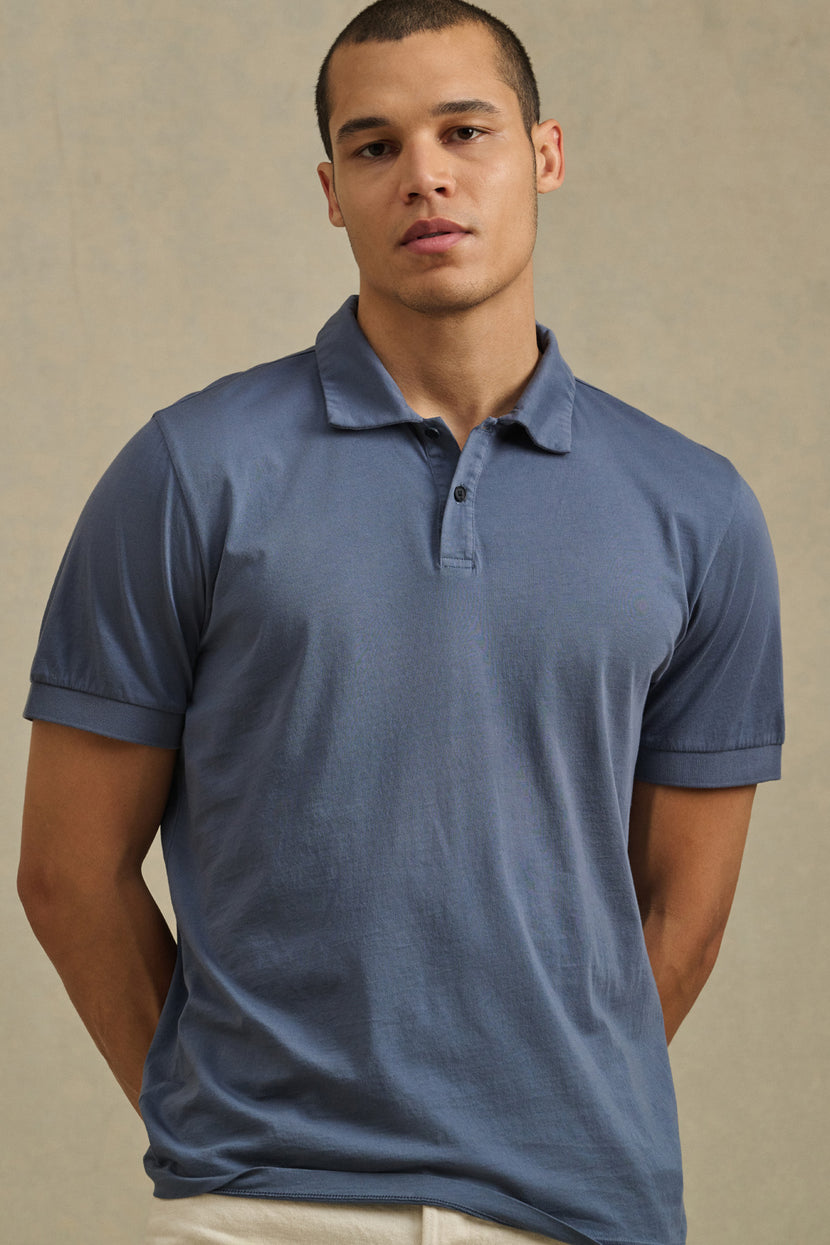 A man wears the ARROW POLO by Velvet by Graham & Spencer, a short-sleeve blue cotton shirt, standing against a beige background and looking directly at the camera for a classic style.