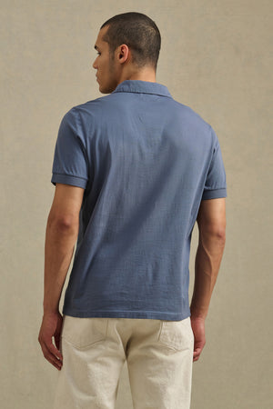 A person faces away wearing the ARROW POLO by Velvet by Graham & Spencer—a classic style, short-sleeve, lightweight blue polo—paired with off-white pants against a plain background.