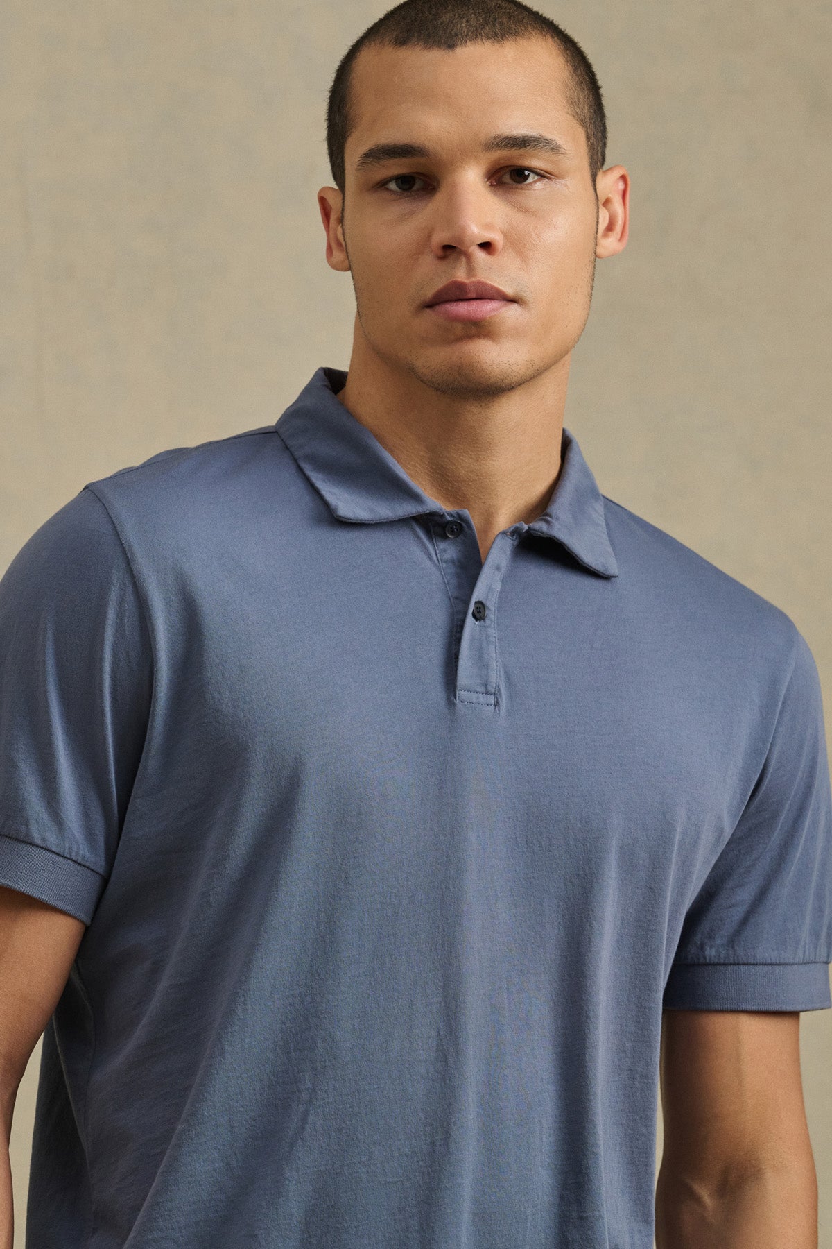 A person models the ARROW POLO by Velvet by Graham & Spencer, a short-sleeved, light blue cotton shirt, standing against a plain beige background to showcase its classic style and comfort.-41104666198209