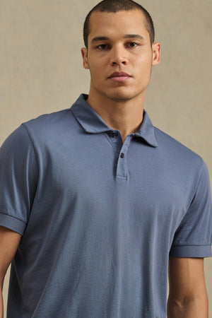 A person models the ARROW POLO by Velvet by Graham & Spencer, a short-sleeved, light blue cotton shirt, standing against a plain beige background to showcase its classic style and comfort.