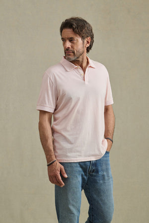 A man wearing the ARROW POLO by Velvet by Graham & Spencer in light pink cotton and blue jeans stands against a beige background, looking to his left.