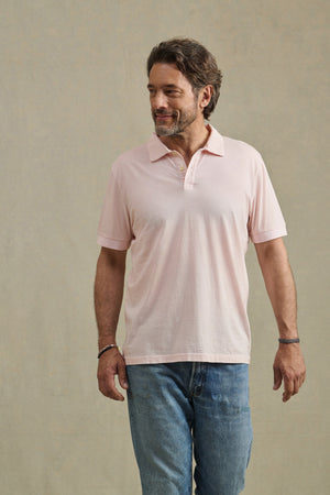 A man with brown hair and a beard is wearing the ARROW POLO by Velvet by Graham & Spencer in light pink with blue jeans, standing against a plain beige background.