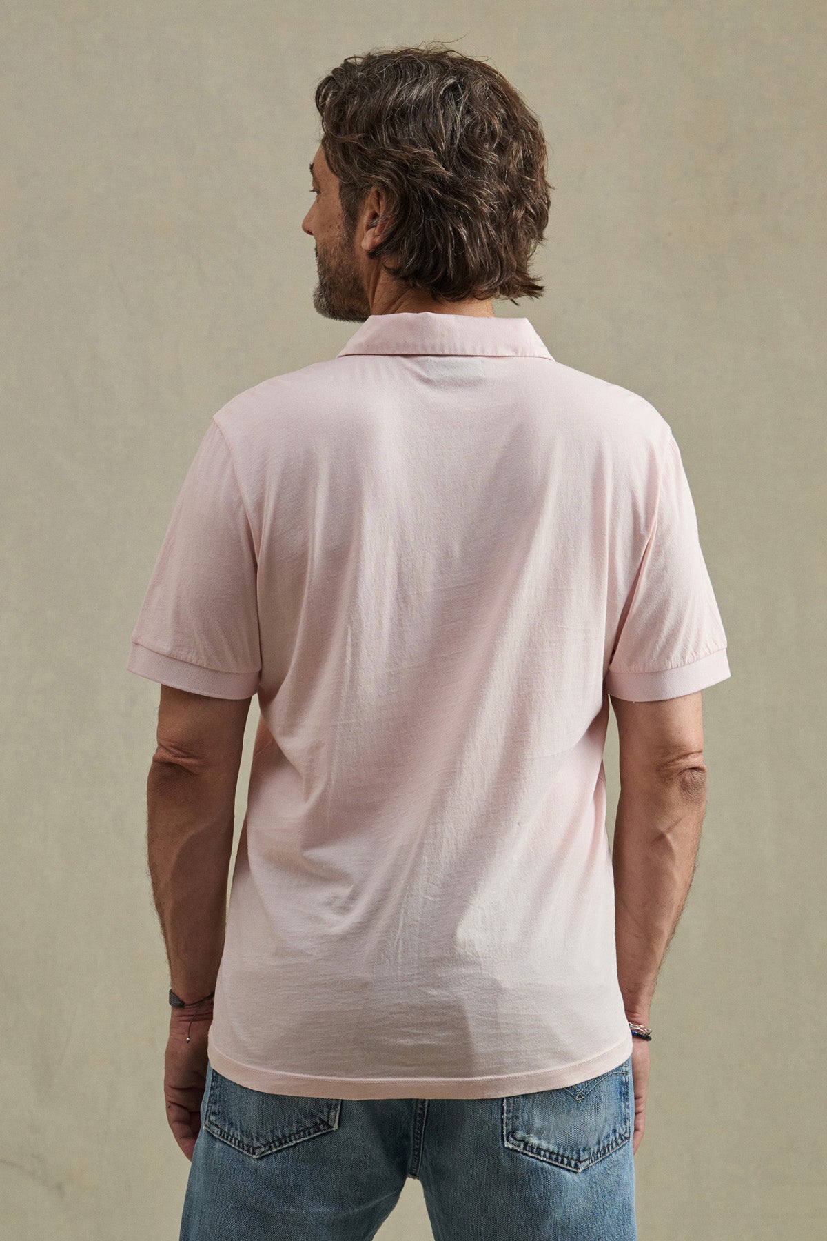 A man faces away from the camera, wearing the ARROW POLO in light pink by Velvet by Graham & Spencer with blue jeans, displaying a classic look against a plain beige backdrop.-41008940777665