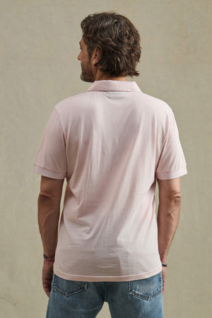 A man faces away from the camera, wearing the ARROW POLO in light pink by Velvet by Graham & Spencer with blue jeans, displaying a classic look against a plain beige backdrop.