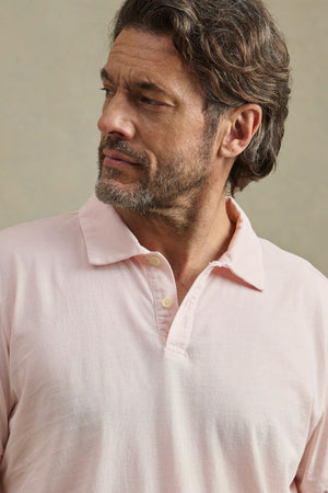 A man with wavy brown hair and a beard wears the ARROW POLO by Velvet by Graham & Spencer in light pink, embracing classic style as he looks to his left against a neutral background.