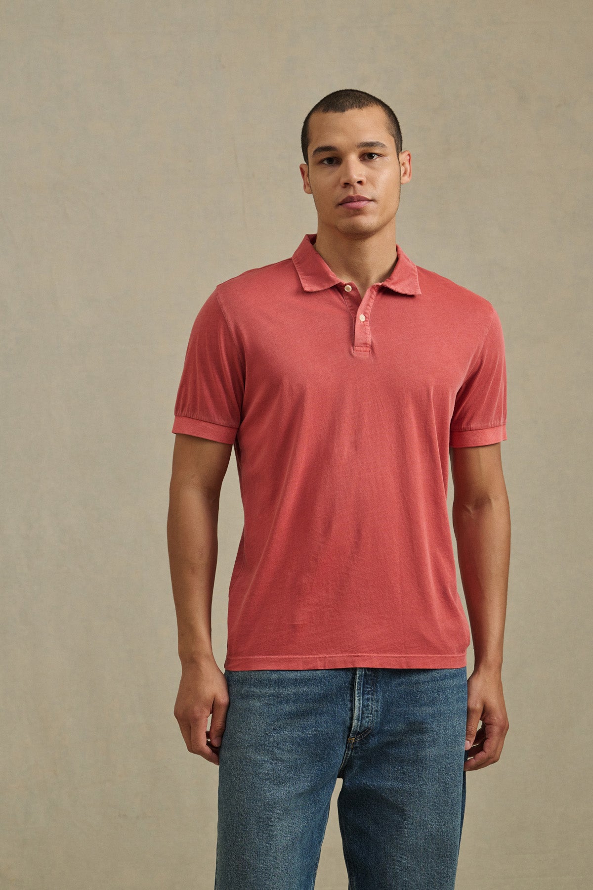 A person wears the ARROW POLO by Velvet by Graham & Spencer, a short-sleeve red casual shirt, paired with blue jeans and standing against a plain light background.