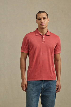 A person wears the ARROW POLO by Velvet by Graham & Spencer, a short-sleeve red casual shirt, paired with blue jeans and standing against a plain light background.